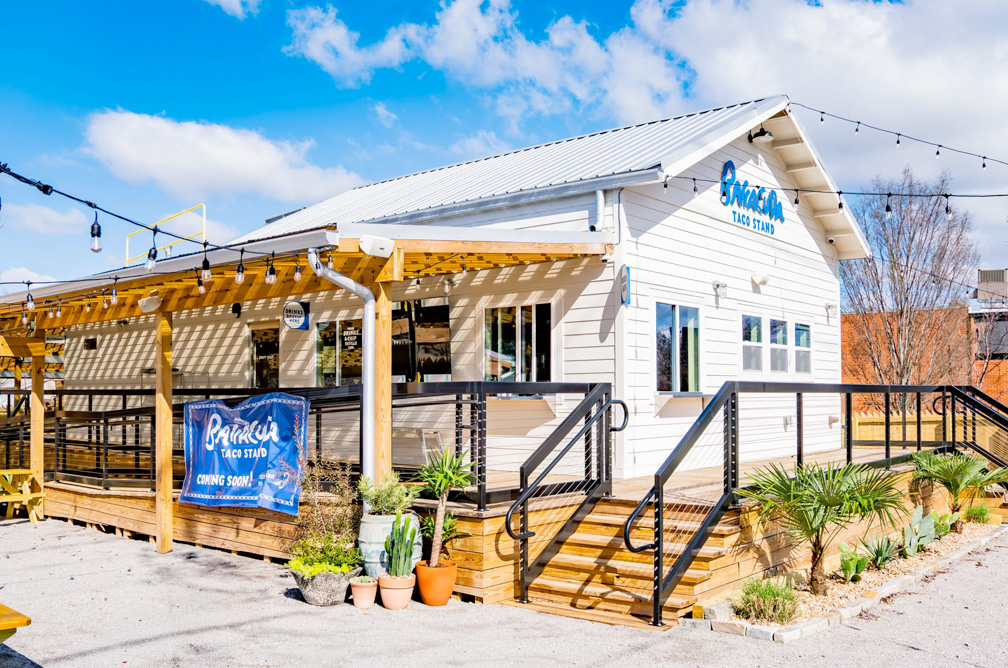 Small white Barracuda taco stand with a wooden pergola and steps, surrounded by potted plants. A "coming soon" sign hangs outside. Bright, sunny day.